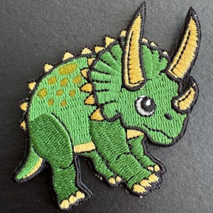 May include: Embroidered patch of a cartoon triceratops dinosaur. The patch features a green body with yellow accents on the horns, frill, and feet. The dinosaur has a friendly expression with a white eye and black outline.