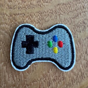 May include: Embroidered patch of a video game controller. The grey controller features a black cross-shaped directional pad and coloured buttons. It has a white border and is set against a wooden background.