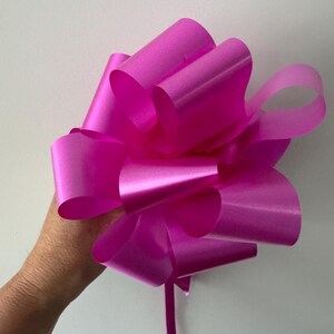 May include: A large, bright pink pull bow made of shiny ribbon. The bow has multiple loops and a long, trailing ribbon. The bow is held in a hand against a plain white background.