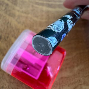 May include: A black pencil with a floral pattern is positioned above a bright pink pencil sharpener. The sharpener is rectangular with a clear lid and a pink interior. The pencil's end is grey.