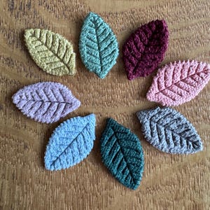 May include: Eight crocheted leaf appliques in a variety of colours, including yellow, teal, burgundy, pink, lavender, blue, green, and grey. The leaves are arranged in a circular pattern on a wooden surface.
