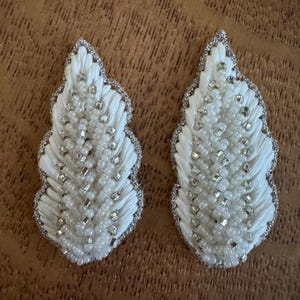 May include: Two white, leaf-shaped embroidered appliques. Each applique features a central design of white thread and small, round white beads, with silver-coloured square and rectangular accents. The edges are trimmed with silver thread.