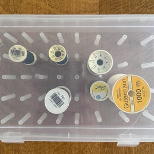 May include: A clear plastic storage box containing several thread spools in shades of white, yellow, and blue. The spools have labels with text and barcodes. The box has pegs to keep the spools organised. Some spools are labelled 'Gütermann'.