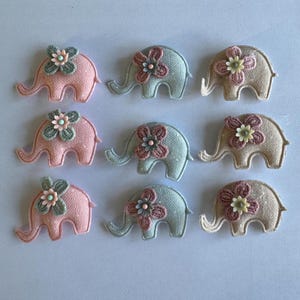 May include: Nine embroidered elephant appliques in pink, blue, and beige. Each elephant has a floral embellishment on its head. The appliques are arranged in three rows of three.