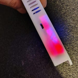 May include: A white rectangular electronic device with a red cable inserted. The device has a series of vents and emits a pink and blue glow. The device is held by a person.