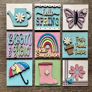 Puede incluir: Nueve letreros de madera con diseños de temática primaveral. Los letreros presentan una regadera, una mariposa, un arcoíris, un paraguas, botas de lluvia, flores y pájaros. Los letreros tienen el texto "hello spring", "bloom & grow", "hello sunshine", "fresh picked daisies".