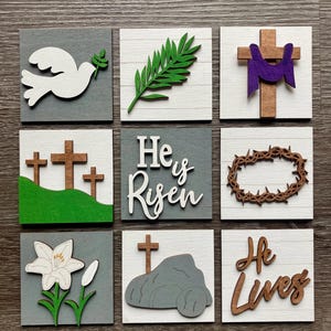 May include: Nine square wooden blocks with Easter-themed designs. The blocks feature a white dove, green palm branch, a cross with purple cloth, three crosses on a green hill, the text "He is Risen", a crown of thorns, a lily, a cross with a stone, and the text "He Lives".