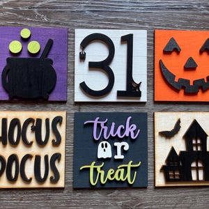 May include: Six wooden signs with Halloween themes. The signs are painted in various colours, including purple, white, orange, black, and brown. The signs feature images and text, including a cauldron, the number 31, a jack-o'-lantern, the words "Hocus Pocus", a ghost, the words "trick or treat", a bat, and a haunted house.