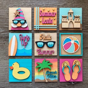 May include: Nine square wooden blocks with summer-themed designs. The blocks feature a gnome, text that says "Summer Lovin'", a sandcastle, a surfboard, sunglasses, a beach ball, a duck float, a palm tree with "Beach Vibes", and flip-flops.