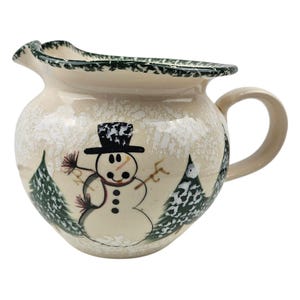 May include: Cream-colored ceramic pitcher with a snowman design. The pitcher features a black top hat, a carrot nose, and a red scarf. Green trees are painted on either side of the snowman. The rim is trimmed with a green, textured pattern.