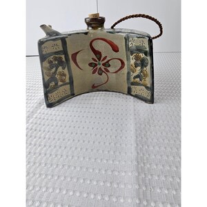 May include: A ceramic flask with a curved shape and a brown woven handle. The flask features a beige background with red and green floral designs. The edges have a dark blue and beige pattern.