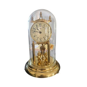 May include: A gold-toned antique clock with a cream-colored face, protected by a clear glass dome. The clock features black Roman numerals and ornate detailing. The base is round and also gold-toned.