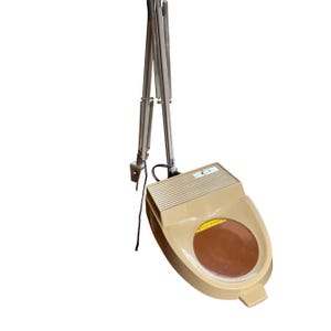 Puede incluir: Una lámpara de aumento beige con una lente grande y circular. La lámpara tiene un brazo articulado flexible y una cabeza rectangular. La lente es marrón y la lámpara está diseñada para trabajos de cerca.