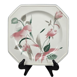May include: An octagonal, off-white plate with a floral design. Pink roses and green leaves are arranged across the plate's surface. The plate has a thin, dark green border. The plate is displayed on a black stand.
