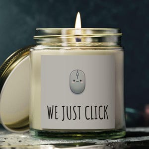 Puede incluir: Una vela en un tarro de vidrio con una etiqueta blanca que dice "WE JUST CLICK" y una imagen de dibujos animados de un ratón de computadora con una cara sonriente.