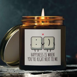 Puede incluir: Una vela de vidrio marrón con una etiqueta blanca que dice "Happiness is when you're right next to me". La etiqueta presenta dos teclas de teclado de dibujos animados tomándose de la mano. Una tecla tiene dos puntos y punto y coma para los ojos y la otra tecla tiene un paréntesis derecho para una sonrisa.