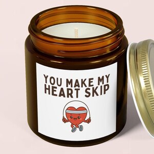 Puede incluir: Una vela en tarro de cristal marrón con una etiqueta blanca que dice "YOU MAKE MY HEART SKIP". La etiqueta presenta un corazón de dibujos animados saltando a la cuerda. La vela tiene una tapa dorada y un interior de cera blanca.