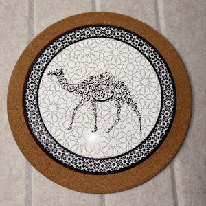 May include: Round coaster with a cork base and a decorative ceramic top. The ceramic features a white background with a geometric pattern and a black and white camel design. A black and white border surrounds the central design.