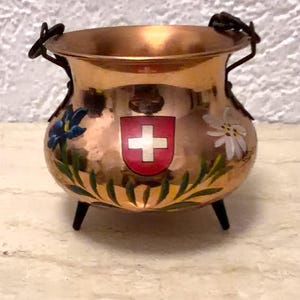 Könnte beinhalten: Ein kleiner, runder, gehämmerter Kupferkessel mit drei schwarzen Beinen und einem schwenkbaren Griff aus dunklem Metall. Die Vorderseite zeigt ein bemaltes rotes Wappenschild mit weißem Kreuz, flankiert von einer blauen Blume und einem Edelweiß.