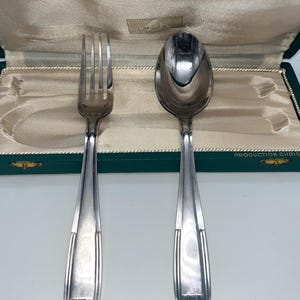 Könnte beinhalten: Ein silbernes Gabel- und Löffelset, ausgestellt außerhalb eines grün samtgefütterten Etuis. Das Besteck hat ein stromlinienförmiges Design mit vertikalen Linien auf den Griffen. Das Etui ist geöffnet und zeigt einen cremefarbenen Innenraum.