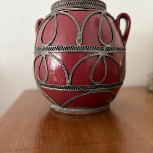 Könnte beinhalten: Eine rote Keramikvase mit silbernen Metallfiligranarbeiten. Die Vase hat eine runde Form, zwei kleine Griffe und einen dekorativen Metallrand. Die Metallarbeiten bilden ein Muster aus verbundenen Tropfenformen.