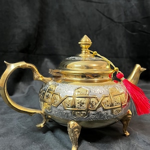 Könnte beinhalten: Verzierte silber-goldene Teekanne mit aufwendigen Mustern und roter Quaste. Die Teekanne hat einen goldenen Griff, Ausguss, Deckel und Füße. Der Korpus ist silberfarben mit goldenen Akzenten. Ein dekoratives Stück für Teeliebhaber.