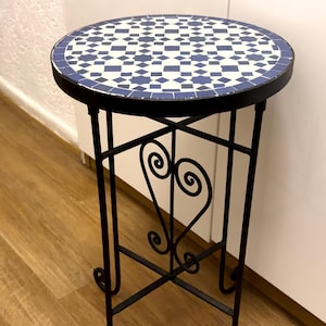 Könnte beinhalten: Ein runder Beistelltisch mit einer blau-weißen, geometrischen Mosaikplatte. Der Tisch wird von einem schwarzen Metallgestell mit vier an der Basis geschwungenen Beinen getragen, wobei dekorative Schnörkel die Beine in der Mitte verbinden.