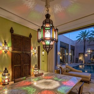 May include: A dining room featuring a large, ornate, multi-colored pendant lamp. The lamp casts colorful light onto a marble table. Additional wall sconces and lanterns add to the warm, inviting ambiance. A large wooden door and a view of an outdoor pool area are also visible.