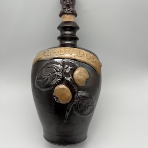 Könnte beinhalten: Eine dunkelbraune Keramikflasche mit einem dekorativen Verschluss mit einem Gesicht. Die Flasche hat ein beiges Banner mit der Aufschrift "CALVADOS". Sie ist mit geprägten Blättern und Früchten verziert. Die Flasche wird wahrscheinlich zur Aufbewahrung von Spirituosen verwendet.