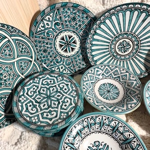 Könnte beinhalten: Eine Sammlung von Keramiktellern und Schalen mit komplizierten, handbemalten Mustern in Türkis, Schwarz und Weiß. Die Designs umfassen geometrische Formen, florale Motive und radiale Linien, wobei jedes Stück eine einzigartige Anordnung zeigt.