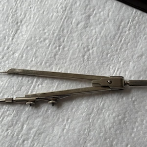 Può includere: Un compasso da disegno in metallo color argento con due bracci incernierati, uno dotato di una punta metallica affilata e l'altro di una mina in grafite. Lo strumento include due piccole rotelle di regolazione e un'impugnatura zigrinata.
