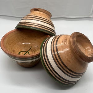Könnte beinhalten: Drei kleine, handgefertigte Keramikschalen. Die Schalen sind beige mit horizontalen Streifen in Schwarz, Weiß und Grün. Eine Schale ist umgedreht und zeigt das Innere, das hellbraun ist und ein grün gemaltes Design aufweist.