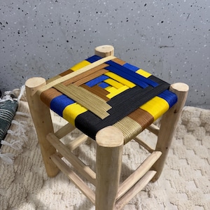 Peut inclure: Un petit tabouret carré en bois avec une assise tissée. L'assise présente un motif coloré de bandes tissées jaunes, bleues, noires et marron. Les pieds sont en bois clair, et le tabouret est conçu pour s'asseoir.