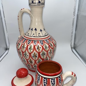 Könnte beinhalten: Ein Keramik-Krug- und Becher-Set mit passendem Deckel. Der Krug hat einen hohen, schlanken Hals und einen abgerundeten Körper mit einem Griff. Das Set ist mit roten, blauen und grünen Mustern auf cremefarbenem Hintergrund bemalt. Der Becher hat einen Griff und ein ähnliches Design.