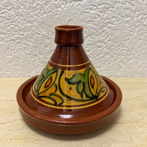 Könnte beinhalten: Eine glasierte Terrakotta-Tajine mit einem konischen Deckel, der einen braunen Boden und einen gelben Mittelteil mit handgemalten grünen und orangen Blumenmotiven aufweist. Das Kochgeschirr besteht aus einer runden Schale und einem Deckel.