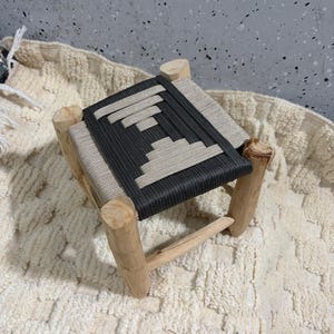 Könnte beinhalten: Kleiner Holzschemel mit gewebter Sitzfläche. Der Sitz hat ein geometrisches Design in Dunkelgrau und Beige. Die Beine sind aus Naturholz gefertigt, und der Hocker steht auf einem strukturierten, cremefarbenen Teppich.