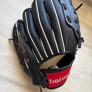 Könnte beinhalten: Ein schwarzer Baseballhandschuh mit weißer Paspelierung entlang der Finger und einem roten rechteckigen Aufnäher am Handgelenkriemen mit dem weißen Schriftzug "barnett". Die Seite des Handschuhs zeigt den goldenen Text "barnettsports.com" und schwarze Schnürung.