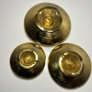 Könnte beinhalten: Drei Aschenbecher aus Messing mit kreisförmigem Design. Jeder Aschenbecher hat eine flache Schalenform mit einer zentralen Vertiefung zum Halten von Zigaretten. Die Aschenbecher sind in einem dreieckigen Muster auf weißem Hintergrund angeordnet.