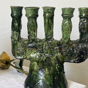 Könnte beinhalten: Ein handgefertigter, grüner Keramik-Menora mit sieben Kerzenhaltern. Die Menora hat ein strukturiertes, organisches Design mit glänzendem Finish. Eine getrocknete gelbe Rose liegt in der Nähe der Basis.