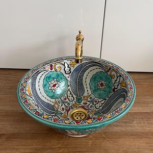 Puede incluir: Lavabo ovalado de cerámica con un colorido diseño floral. El lavabo tiene un grifo y un desagüe dorados. El interior tiene un patrón floral turquesa y blanco con detalles de hojas azules y verdes. El borde está decorado con un patrón repetido de rojo, amarillo y verde.