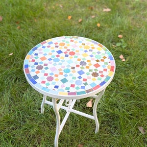 Peut inclure: Une table d'appoint ronde dotée d'un plateau en mosaïque avec un motif géométrique composé de petites tuiles multicolores dans des tons de bleu, rouge, jaune, vert et blanc. La table possède un cadre en métal blanc avec quatre pieds incurvés.