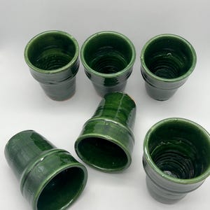 Puede incluir: Seis macetas de cerámica verdes con acabado brillante. Las macetas tienen forma cilíndrica con un borde ligeramente ensanchado y una textura acanalada. Las macetas están dispuestas sobre una superficie blanca.