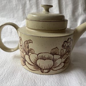Könnte beinhalten: Eine cremefarbene Keramik-Teekanne mit gesprenkelter Oberfläche und einem braunen Blumenmuster als Strichzeichnung. Die Teekanne hat einen zylindrischen Körper, einen geschwungenen Henkel, einen kurzen Ausguss und einen passenden Deckel.
