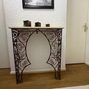 Könnte beinhalten: Eine dekorative Konsole mit einer hellfarbigen Steinplatte und einem verzierten, dunkelbraunen Schmiedeeisenfuß. Der Fuß hat ein sich wiederholendes Scroll-Design. Drei kleine dekorative Boxen stehen auf dem Tisch. Ein gerahmtes Schwarz-Weiß-Bild hängt über dem Tisch.