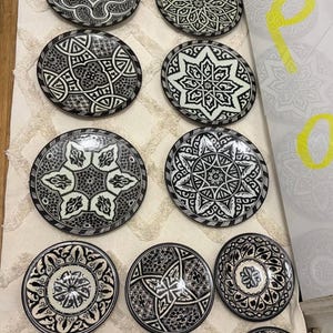Könnte beinhalten: Eine Sammlung von vierzehn runden Keramiktellern mit komplizierten schwarz-weißen geometrischen und floralen Mustern. Die Teller variieren in der Größe und zeigen verschiedene symmetrische Designs, darunter sternförmige Motive, verschlungene Linien und verzierte Ränder.