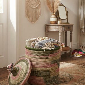 May include: A cylindrical woven storage basket with a removable lid, featuring a multi-colored pattern of olive green, soft pink, and natural tan fibers. The basket is filled with several folded textile blankets with fringed edges.