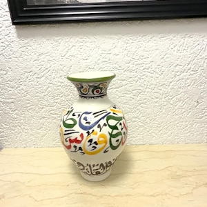 Könnte beinhalten: Eine weiße Keramikvase mit ausgestelltem Rand und bauchigem Körper, verziert mit handgemalter arabischer Kalligrafie in Rot, Blau, Gelb und Grün. Der Rand ist mit einem dünnen grünen Streifen versehen und der Fuß verjüngt sich.