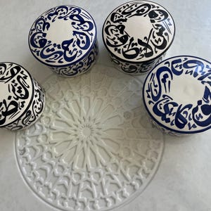 Könnte beinhalten: Vier Keramik-Schmuckdosen mit Deckel. Die Dosen sind weiß mit blauen und schwarzen Kalligraphie-Designs. Die Ränder sind blau. In der Mitte befindet sich ein weißes, dekoratives, kreisförmiges Tablett.