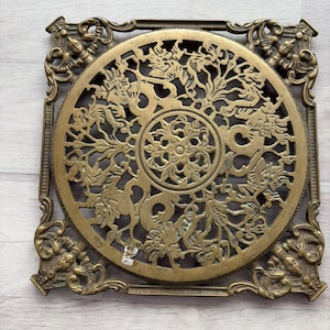 May include: A square brass trivet featuring an intricate openwork design. The center displays a circular medallion with a floral motif, surrounded by stylized dragon figures. The four corners are decorated with ornate, winged cherub-like figures.