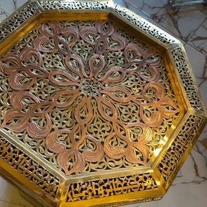 Könnte beinhalten: Dekorativer achteckiger Tisch mit detailliertem, durchbrochenem Design. Der Tisch hat einen Messingrahmen und eine Oberseite aus Kupfer- und Messingfiligran. Das komplizierte Muster ist mit einem floralen Motiv zentriert. Ein einzigartiges Wohnaccessoire, ca. 60 cm Durchmesser.